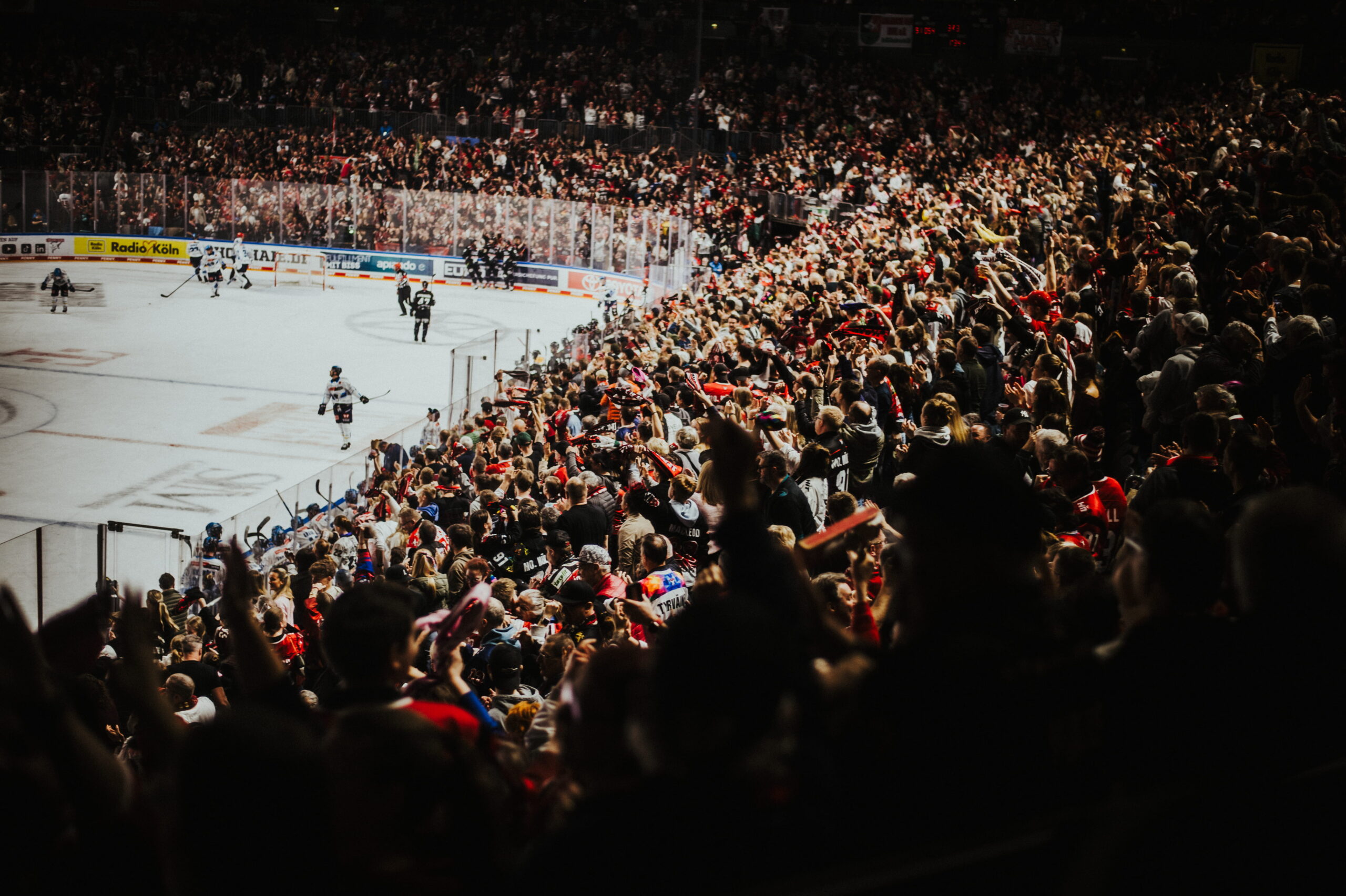 Arena der Kölner Haie. Links im Bild das Spielfeld mit einigen Spielern, rechts im Bild die vollen Besucherränge.
